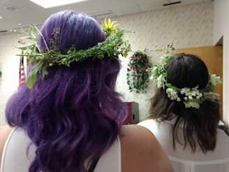 Two individuals with floral crowns are seen from behind, standing in what appears to be an indoor room with neutral wallpaper and some floral decorations. One person has long, wavy purple hair and the other has brown hair, both with intricate crowns of greenery and small white flowers.