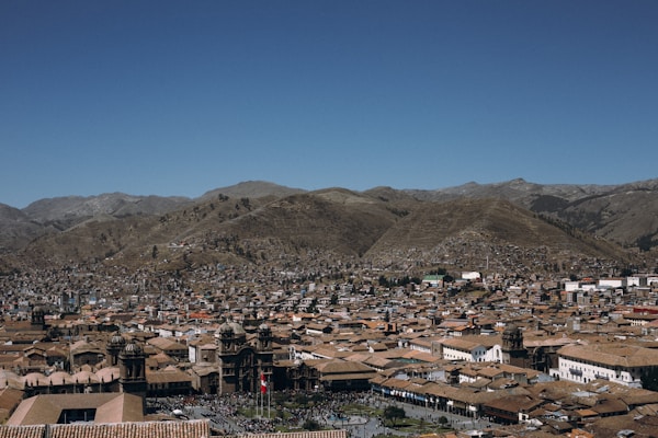 A panoramic view of a city surrounded by mountains. The city is densely packed with traditional buildings featuring red-tiled roofs. Prominent structures, including a cathedral and other significant buildings, are visible in the foreground. The sky is clear with a deep blue hue, and the mountains in the background appear brown and rugged.