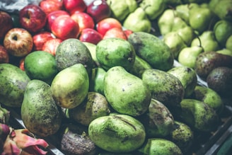 A market display features an assortment of fruits including green avocados, shiny red apples, and light green pears. The fruits are piled together, showcasing their natural textures and variety.