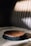 A round chocolate cake with a smooth, glossy surface sits on a patterned fabric surface. Sunlight casts soft shadows across the cake, enhancing its rich texture. The background features stripes with a slightly blurred effect, creating a cozy and intimate atmosphere.