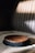 A round chocolate cake with a smooth, glossy surface sits on a patterned fabric surface. Sunlight casts soft shadows across the cake, enhancing its rich texture. The background features stripes with a slightly blurred effect, creating a cozy and intimate atmosphere.