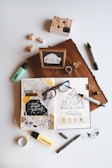 An artistic workspace featuring an open journal with creative doodles, mood tracker, and inspirational quotes. Around the journal, there are markers, a box of alphabet stamps, loose stamps, a wooden block with a framed quote, glasses, tape, and small decorative pieces. The setup is organized on a light background.