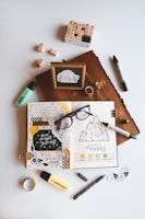 An artistic workspace featuring an open journal with creative doodles, mood tracker, and inspirational quotes. Around the journal, there are markers, a box of alphabet stamps, loose stamps, a wooden block with a framed quote, glasses, tape, and small decorative pieces. The setup is organized on a light background.