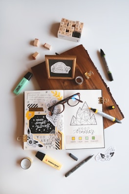 An artistic workspace featuring an open journal with creative doodles, mood tracker, and inspirational quotes. Around the journal, there are markers, a box of alphabet stamps, loose stamps, a wooden block with a framed quote, glasses, tape, and small decorative pieces. The setup is organized on a light background.
