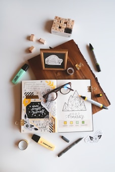 An artistic workspace featuring an open journal with creative doodles, mood tracker, and inspirational quotes. Around the journal, there are markers, a box of alphabet stamps, loose stamps, a wooden block with a framed quote, glasses, tape, and small decorative pieces. The setup is organized on a light background.