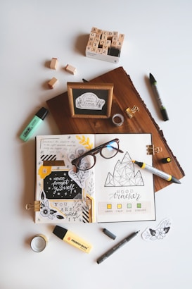 An artistic workspace featuring an open journal with creative doodles, mood tracker, and inspirational quotes. Around the journal, there are markers, a box of alphabet stamps, loose stamps, a wooden block with a framed quote, glasses, tape, and small decorative pieces. The setup is organized on a light background.