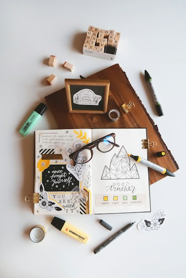 An artistic workspace featuring an open journal with creative doodles, mood tracker, and inspirational quotes. Around the journal, there are markers, a box of alphabet stamps, loose stamps, a wooden block with a framed quote, glasses, tape, and small decorative pieces. The setup is organized on a light background.