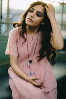 A woman with long, wavy hair is seated in a relaxed pose, gently touching her hair with one hand and holding a meditative position with the other. She is wearing a soft pink dress and a necklace with large pendants. The background is out of focus, suggesting an urban setting with a fence.