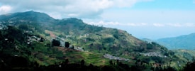 A panoramic view of lush green hills with terraced fields sprawling across the landscape, dotted with small clusters of houses. The hills are enveloped by a partly cloudy sky, creating a serene rural vista.