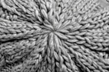 A close-up view of a knitted fabric displaying a textured pattern of interwoven yarns. The fibers are thick, creating a pronounced and intricate design with a star-shaped center.