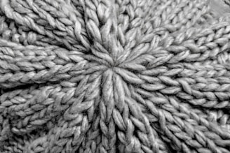 A close-up view of a knitted fabric displaying a textured pattern of interwoven yarns. The fibers are thick, creating a pronounced and intricate design with a star-shaped center.