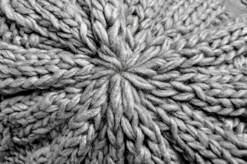 A close-up view of a knitted fabric displaying a textured pattern of interwoven yarns. The fibers are thick, creating a pronounced and intricate design with a star-shaped center.