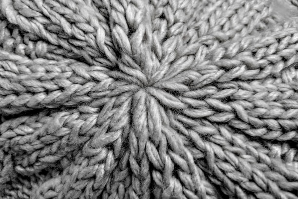 A close-up view of a knitted fabric displaying a textured pattern of interwoven yarns. The fibers are thick, creating a pronounced and intricate design with a star-shaped center.