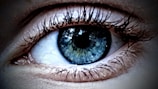 A close-up view of a human eye with intricate details of the iris and eyelashes visible. The iris displays a mixture of blue and green tones, with reflections creating a glossy effect. Eyelashes are sharply defined and frame the eye prominently.