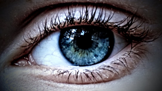 A close-up view of a human eye with intricate details of the iris and eyelashes visible. The iris displays a mixture of blue and green tones, with reflections creating a glossy effect. Eyelashes are sharply defined and frame the eye prominently.