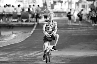 An elderly person is riding a bicycle on a paved path. They appear relaxed, surrounded by several people standing and walking in the background near a railing beside a body of water. The pathway and trees create a serene atmosphere.
