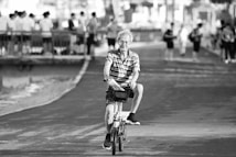 An elderly person is riding a bicycle on a paved path. They appear relaxed, surrounded by several people standing and walking in the background near a railing beside a body of water. The pathway and trees create a serene atmosphere.