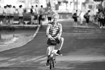An elderly person is riding a bicycle on a paved path. They appear relaxed, surrounded by several people standing and walking in the background near a railing beside a body of water. The pathway and trees create a serene atmosphere.