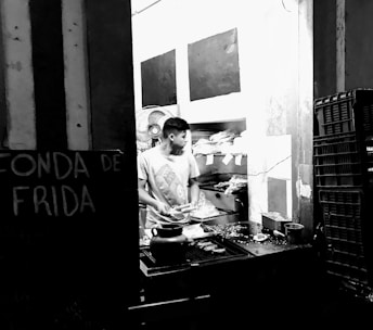 A person is standing in a small food preparation area, likely a street food stall or small kitchen. They are engaged in preparing or handling food. There is a visible sign on the left that partially reads 'FONDA DE FRIDA'. The environment is cluttered with cooking utensils, and there are stacks of containers on the right side.