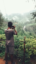 A person standing on a viewing platform or path is taking a photograph of a lush, green landscape. The scenery includes dense vegetation, a distant waterfall cascading down a mountain, and mist or low clouds hanging in the air. The setting appears tranquil and is surrounded by nature.