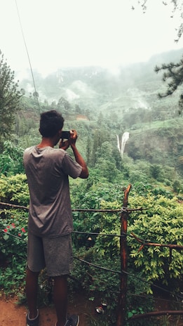 A person standing on a viewing platform or path is taking a photograph of a lush, green landscape. The scenery includes dense vegetation, a distant waterfall cascading down a mountain, and mist or low clouds hanging in the air. The setting appears tranquil and is surrounded by nature.