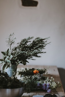 A rustic table is adorned with a variety of green plants and herbs, including rosemary and sage. Some bright orange flowers provide contrast, while other dried herbs are spread around. The wooden surface and muted background give a natural, earthy feel to the setting.
