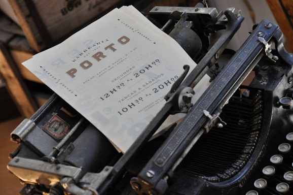 An old-fashioned typewriter holds a sheet of paper with printed text. The paper includes the word 'PORTO' prominently and appears to be some form of advertisement or notice, with additional information about hours or scheduling.