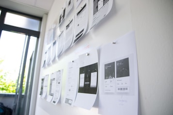 A series of printed pages with design layouts are pinned to a whiteboard in a brightly lit room. The pages appear to be drafts or wireframes of web or app designs, featuring various text and graphical elements. Sunlight filters through nearby windows, creating a mix of natural and artificial lighting.