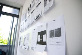 A series of printed pages with design layouts are pinned to a whiteboard in a brightly lit room. The pages appear to be drafts or wireframes of web or app designs, featuring various text and graphical elements. Sunlight filters through nearby windows, creating a mix of natural and artificial lighting.