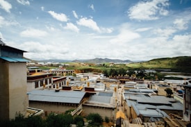 A scenic view of a village with traditional architectural roofs, nestled amidst green fields and distant rolling hills under a partly cloudy sky. The village features several low-rise buildings with distinct designs, surrounded by lush greenery and water bodies in the background.