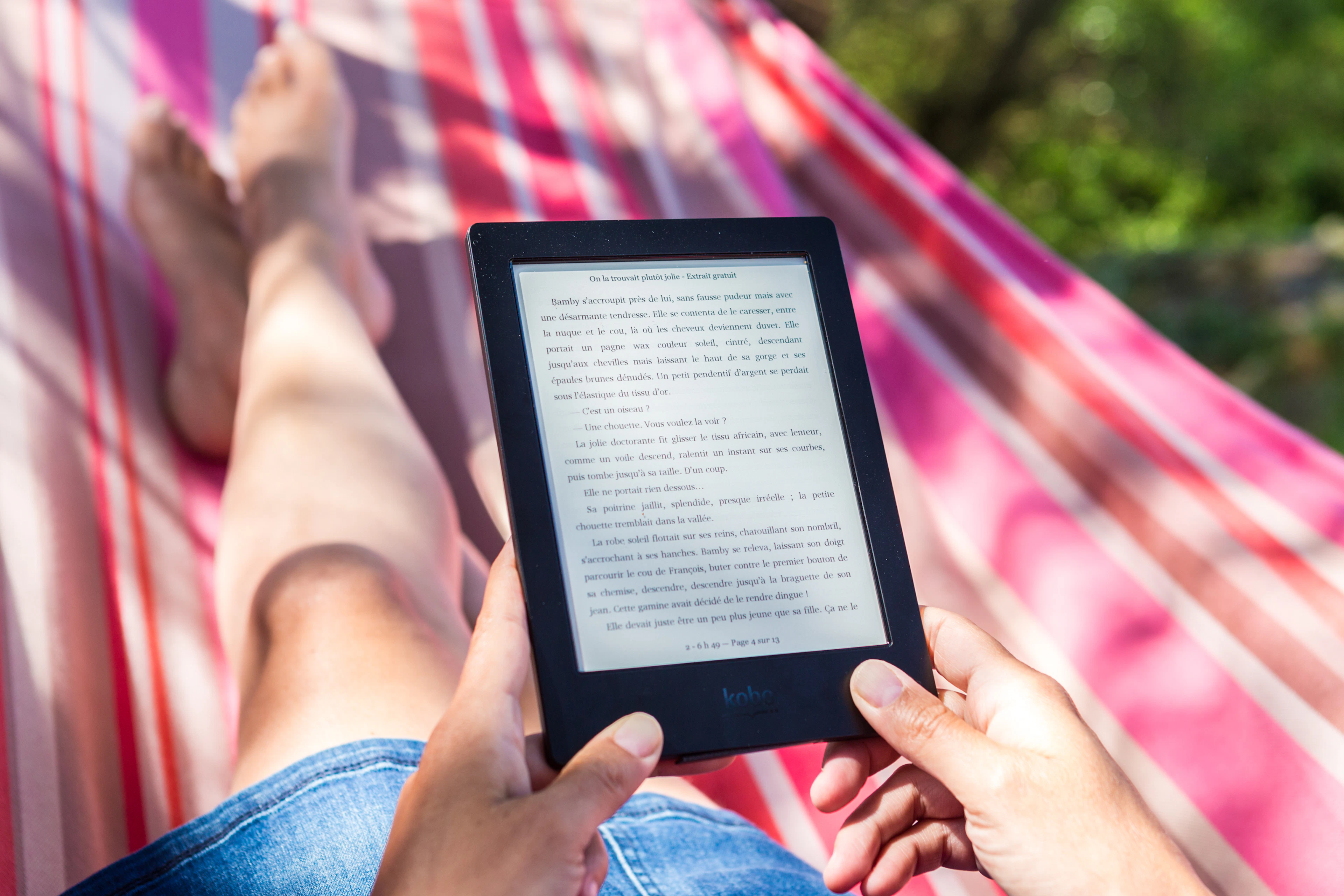 Two hands holding an e-reader above a person's legs stretched out in a red and white striped hammock