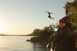 person diving from cliff to body of water during sunset