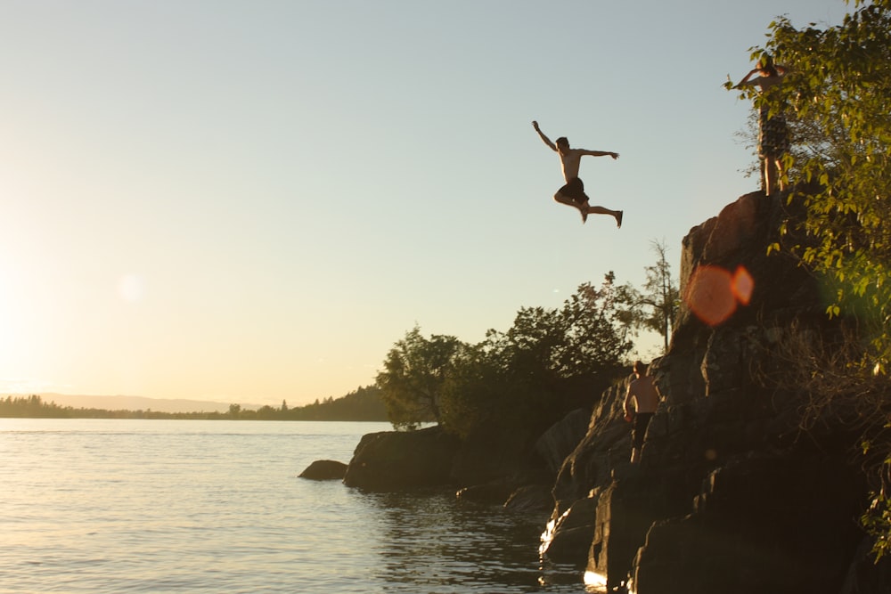 person diving from cliff to body of water during sunset