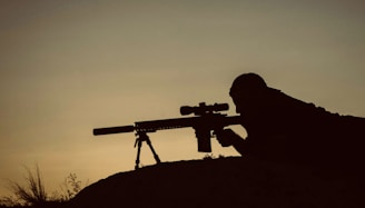 Silhouette of a person lying prone on a hill, aiming a sniper rifle equipped with a scope and bipod. The foreground is in shadow, emphasizing the outline of the rifle and the person. The background is a gradient of warm colors, suggesting sunset or sunrise.
