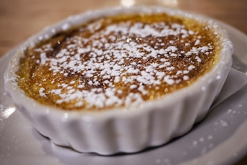 A close-up view of a dessert served in a white ceramic ramekin. The dish appears to be cr&egrave;me br&ucirc;l&eacute;e with a caramelized sugar crust on top, sprinkled with powdered sugar. The golden brown topping contrasts with the white ramekin and plate.