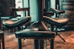 A close-up view of several leather bar stools with metal supports, arranged around a central pillar. The background includes blurred wooden elements, indicating a rustic interior setting with warm lighting.
