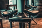 A close-up view of several leather bar stools with metal supports, arranged around a central pillar. The background includes blurred wooden elements, indicating a rustic interior setting with warm lighting.