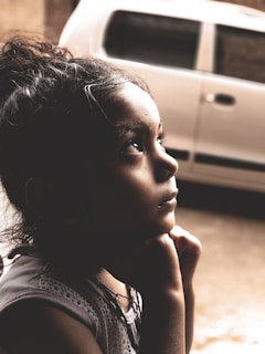 A young child is gazing upwards with a thoughtful expression, hands resting under the chin. The child is positioned in front of a blurred background, which features part of a parked car.