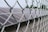 A close-up view of a metal chain-link fence with solar panels visible in the background, set against a slightly overcast sky. The focus is on the fence while the solar panels are slightly blurred.