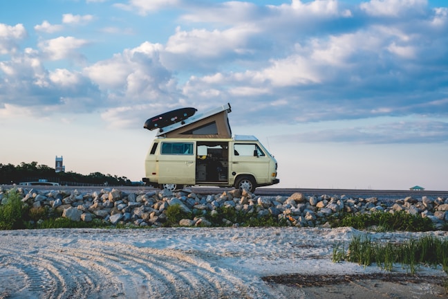 A yellow camper van with a roof tent is parked on a rocky shoreline. There are some grassy patches nearby and a partly cloudy blue sky in the background. A surfboard is mounted on top of the van.