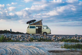 A yellow camper van with a roof tent is parked on a rocky shoreline. There are some grassy patches nearby and a partly cloudy blue sky in the background. A surfboard is mounted on top of the van.