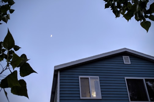 A blue house with visible siding is partially obscured by leafy branches in the foreground. The sky is clear with a crescent moon positioned in the upper left corner.