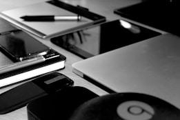 A black and white image featuring various electronic devices and office supplies arranged neatly on a wooden surface. Visible items include a laptop, tablet, smartphone, notebook, pen, and a wireless mouse.