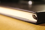 A close-up view of the edge of a closed hardcover book resting on a wooden surface. The leather-bound cover appears slightly worn, and a braided brown cord bookmark is partially visible peeking out from the pages.