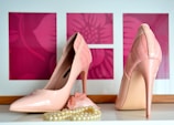 A pair of glossy, pink high-heeled shoes is displayed on a white surface. In front of the shoes, there is a strand of cream-colored pearls partially wrapped around a small pink fabric accessory. The background consists of three pink rectangles featuring floral patterns.