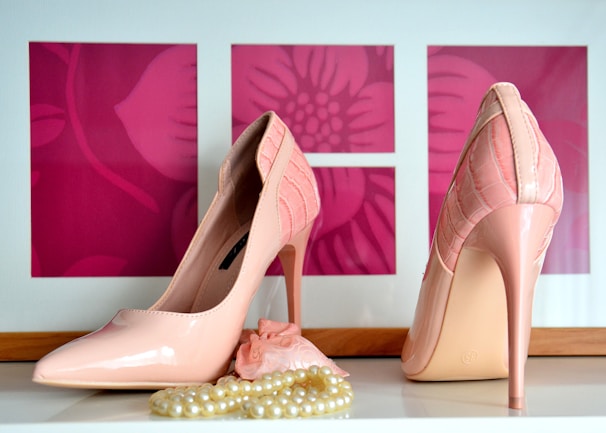 A pair of glossy, pink high-heeled shoes is displayed on a white surface. In front of the shoes, there is a strand of cream-colored pearls partially wrapped around a small pink fabric accessory. The background consists of three pink rectangles featuring floral patterns.