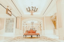A luxurious interior setting featuring an ornate chandelier hanging from a high ceiling, with elegant architectural details such as decorative columns and arches. The center of the image has a vintage-style, red upholstered settee placed on an intricate patterned carpet.