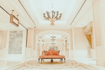 A luxurious interior setting featuring an ornate chandelier hanging from a high ceiling, with elegant architectural details such as decorative columns and arches. The center of the image has a vintage-style, red upholstered settee placed on an intricate patterned carpet.
