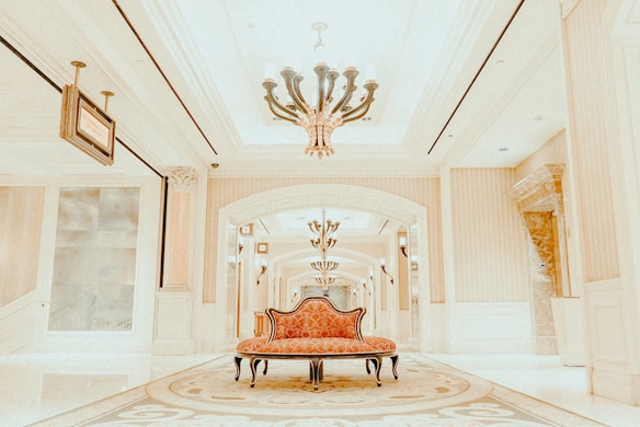 A luxurious interior setting featuring an ornate chandelier hanging from a high ceiling, with elegant architectural details such as decorative columns and arches. The center of the image has a vintage-style, red upholstered settee placed on an intricate patterned carpet.