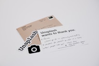 A white card with a handwritten message and printed text reads 'Unsplash wants to thank you.' The card is placed on a table with a brown envelope and Unsplash stickers. The message on the card expresses gratitude, and the envelope has an address label.